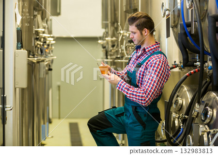 Male brewer in overalls kneeling by brewery tanks examining glass of pale amber beer, copy space 132983813