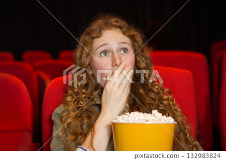 Woman holding yellow popcorn bucket and gasping at screen in dim theater with red upholstered seats Woman holding yellow popcorn bucket and gasping at screen in dim theater with red upholstered seats 132983824