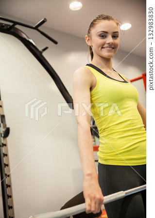 Woman wearing yellow tank top and black leggings, holding metal weight bar at waist in gym 132983830