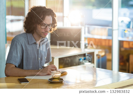Adult man wearing glasses writing in notebook at coffee shop counter with coffee cup and phone 132983832