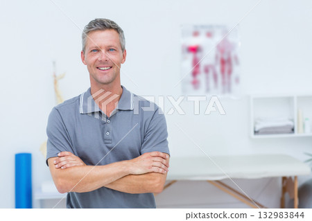 Mature male therapist standing arms crossed in therapy room next to treatment table Mature male therapist standing arms crossed in therapy room next to treatment table 132983844