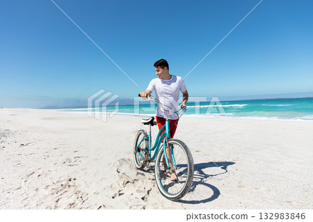Man pushing teal bicycle along white sand beach under clear blue sky with turquoise ocean horizon 132983846
