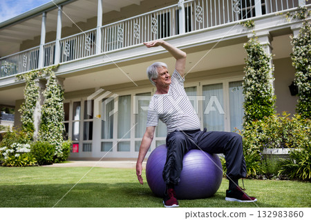 Senior man balancing on purple exercise ball and leaning sideways on lawn by two-story building Senior man balancing on purple exercise ball and leaning sideways on lawn by two-story building 132983860