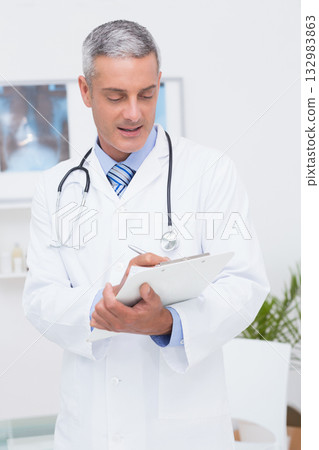 Male doctor writing on clipboard in clinic room with stethoscope and X-ray lightbox Male doctor writing on clipboard in clinic room with stethoscope and X-ray lightbox 132983863