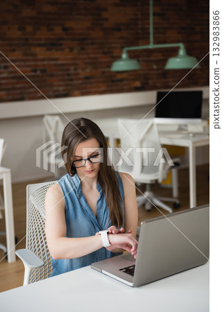 Woman in denim top checking smartwatch while sitting at desk with open laptop in coworking space 132983866