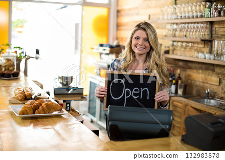 Blonde woman standing behind counter in bakery cafe holding Open chalkboard sign with croissants 132983878