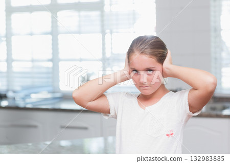 Female child standing at kitchen countertop pressing hands to ears under window with white blinds Female child standing at kitchen countertop pressing hands to ears under window with white blinds 132983885
