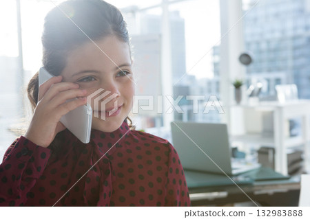 Child in polka dot blouse holding smartphone at office window by desk with laptop, copy space 132983888