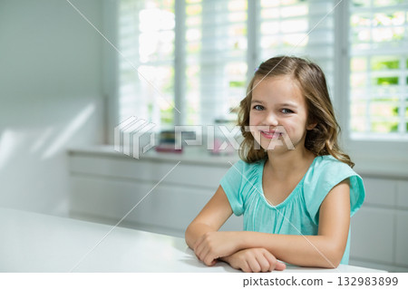 Child girl sitting at white tabletop in bright modern kitchen, smiling under window shutters 132983899