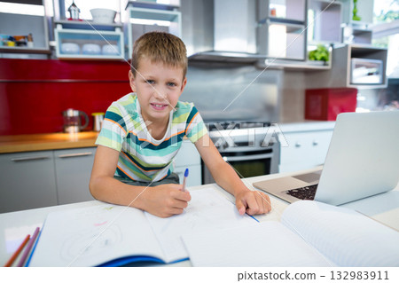 School-age boy writing in notebook with blue pen at kitchen counter with laptop, colored pencils School-age boy writing in notebook with blue pen at kitchen counter with laptop, colored pencils 132983911