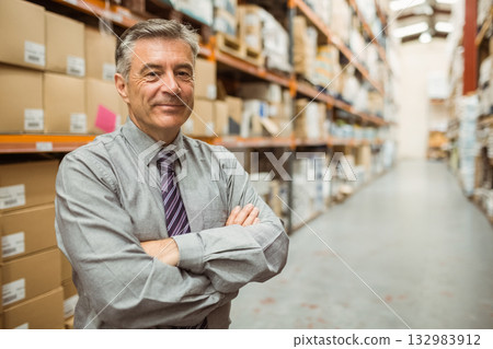 Middle-aged man standing in warehouse wearing business attire with boxes under fluorescent lights 132983912
