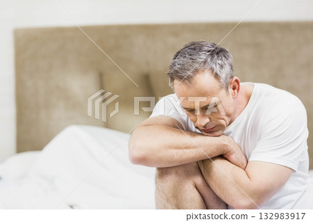 Middle-aged man sitting on bed holding knees in bedroom with white sheets and beige headboard Middle-aged man sitting on bed holding knees in bedroom with white sheets and beige headboard 132983917
