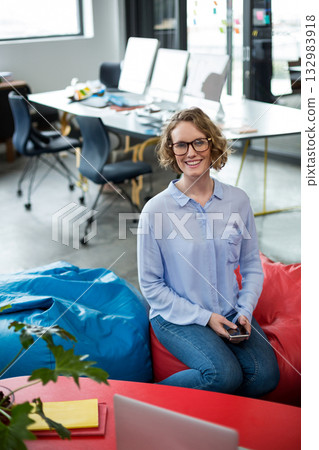 White woman in twenties using smartphone while sitting on beanbag chairs in open office with laptop 132983918