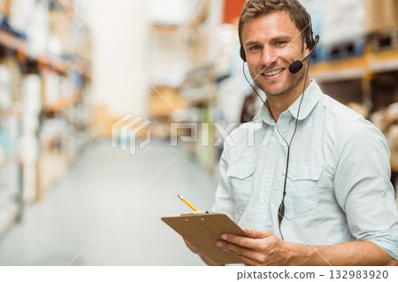 Man standing in warehouse aisle holding clipboard and pencil wearing headset and checking shelves 132983920