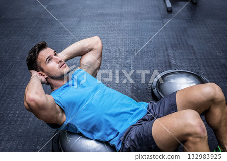Man performing abdominal crunches on half-sphere BOSU trainer at gym with weight machines Man performing abdominal crunches on half-sphere BOSU trainer at gym with weight machines 132983925