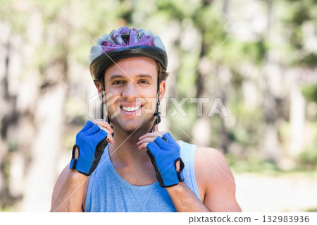 Man adjusting bicycle helmet straps while standing in wooded park wearing blue cycling gloves 132983936