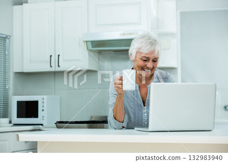 Senior woman standing behind island in home kitchen holding coffee mug and looking at silver laptop Senior woman standing behind island in home kitchen holding coffee mug and looking at silver laptop 132983940