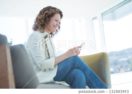 Senior woman sitting on sofa with floor-to-ceiling windows at home holding smartphone and smiling 132983953