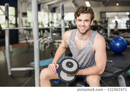 Fit man lifting dumbbell while sitting on weight bench at gym with squat rack stability ball 132983971