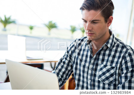Man working at table in café on silver laptop near windows with potted palms, copy space 132983984