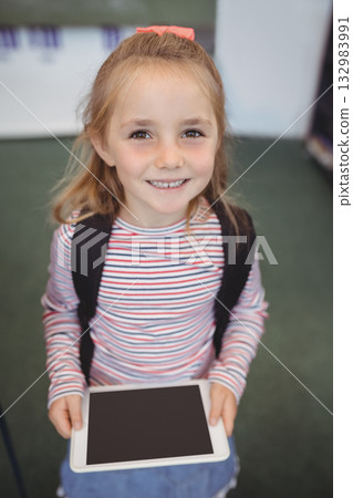 Female child standing on muted green carpet inside hall holding tablet and wearing black backpack Female child standing on muted green carpet inside hall holding tablet and wearing black backpack 132983991