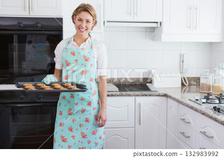 Woman holding cookie tray in kitchen by double oven wearing pastel floral apron and oven mitt Woman holding cookie tray in kitchen by double oven wearing pastel floral apron and oven mitt 132983992