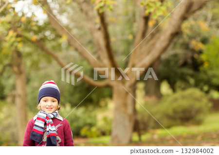 Boy standing, smiling by tree trunk in autumn park, wearing striped knit hat, scarf and sweater Boy standing, smiling by tree trunk in autumn park, wearing striped knit hat, scarf and sweater 132984002