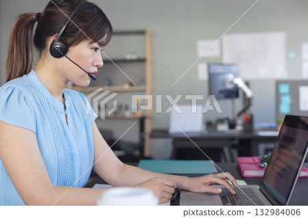 Asian woman speaking into headset and typing on laptop at office desk with coffee cup Asian woman speaking into headset and typing on laptop at office desk with coffee cup 132984006