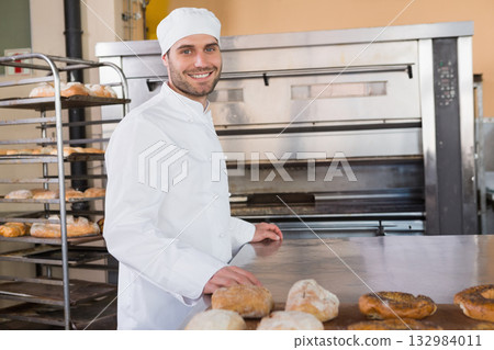 Male baker wearing chef coat smiling while holding fresh loaf behind countertop with rack and oven 132984011