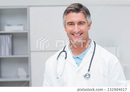 Middle-aged male doctor in lab coat standing in clinic in front of shelving unit with stethoscope 132984017