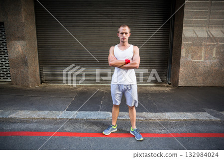 Man wearing athletic clothing standing with arms crossed behind red painted line on narrow street 132984024