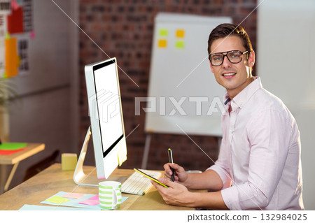 Male in striped shirt writing notes on yellow notepad at office desk with computer, sticky notes 132984025
