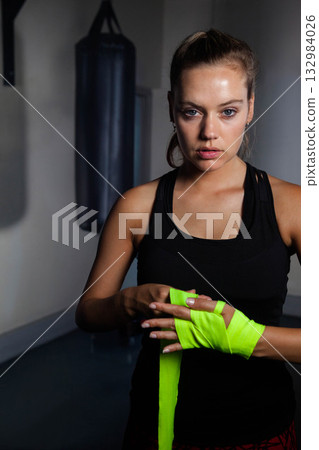 Woman wrapping bright green hand wraps on knuckles beside punching bag at boxing gym, copy space Woman wrapping bright green hand wraps on knuckles beside punching bag at boxing gym, copy space 132984026