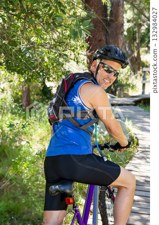 Man sitting on purple mountain bike wearing helmet with hydration pack on boardwalk sunlit forest 132984027