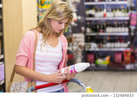 Female shopper leaning on cart examining bottle label carrying bag in supermarket aisle, copy space Female shopper leaning on cart examining bottle label carrying bag in supermarket aisle, copy space 132984033