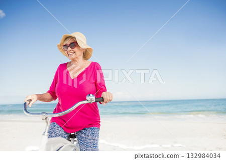 Senior woman standing on beach wearing straw sun hat and sunglasses holding white cruiser bicycle Senior woman standing on beach wearing straw sun hat and sunglasses holding white cruiser bicycle 132984034