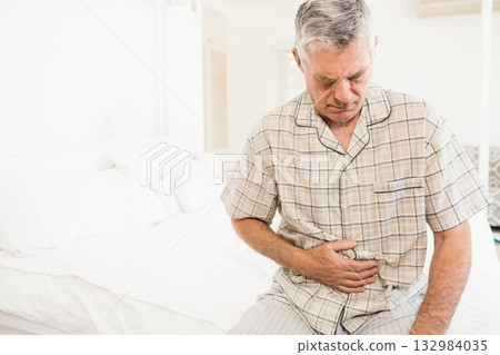 Senior man in checkered pajamas sitting on bed pressing hand on abdomen in bedroom, copy space Senior man in checkered pajamas sitting on bed pressing hand on abdomen in bedroom, copy space 132984035
