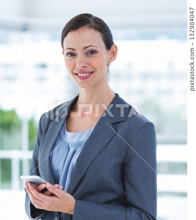 Woman wearing grey blazer and light blue blouse, holding smartphone while smiling in office lobby Woman wearing grey blazer and light blue blouse, holding smartphone while smiling in office lobby 132984047