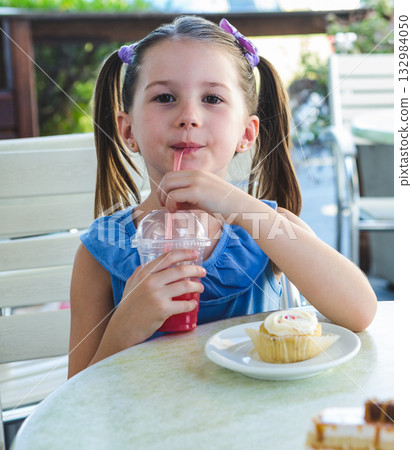 Girl sitting on cafe patio sipping bright red drink through plastic cup and nibbling cupcake Girl sitting on cafe patio sipping bright red drink through plastic cup and nibbling cupcake 132984050