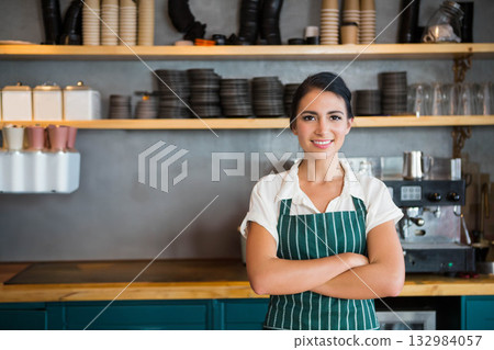 Female barista wearing green-striped apron standing behind counter in cafe using espresso machine 132984057