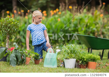Girl child standing with foot resting on potted plants near watering can and wheelbarrow in garden 132984074