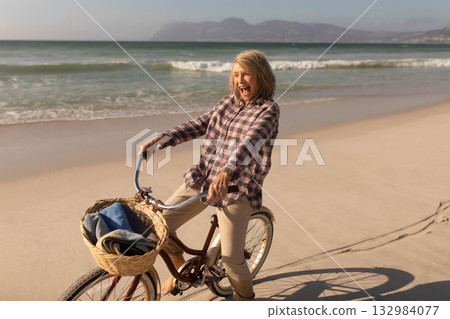 Senior woman wearing checkered shirt riding bicycle on beach with blanket in wicker basket laughing Senior woman wearing checkered shirt riding bicycle on beach with blanket in wicker basket laughing 132984077