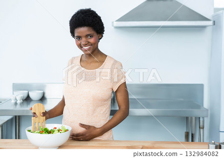 African American woman standing behind kitchen countertop, stirring salad in white bowl with server African American woman standing behind kitchen countertop, stirring salad in white bowl with server 132984082