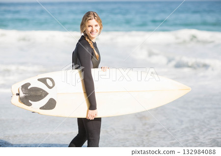 Adult female surfer in black wetsuit standing on shore holding surfboard facing ocean, copy space 132984088