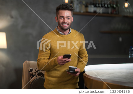 Man wearing mustard sweater holding smartphone and coffee cup at cafe counter near wooden shelves Man wearing mustard sweater holding smartphone and coffee cup at cafe counter near wooden shelves 132984186