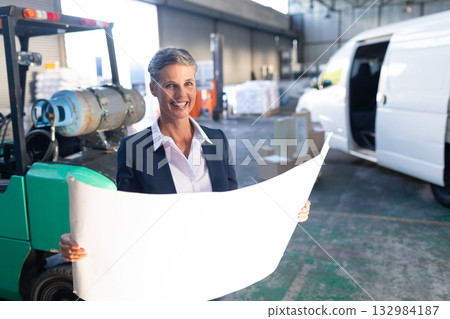 Middle-aged woman wearing blazer at warehouse holding blueprint near green forklift and cargo van Middle-aged woman wearing blazer at warehouse holding blueprint near green forklift and cargo van 132984187