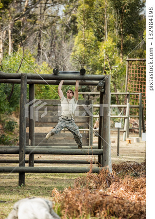 Male soldier swinging from log above beams in forest obstacle course wearing camouflage uniform Male soldier swinging from log above beams in forest obstacle course wearing camouflage uniform 132984198