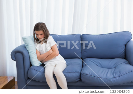 Girl hugging light-blue throw pillow on sofa in living room beside wooden side table, copy space Girl hugging light-blue throw pillow on sofa in living room beside wooden side table, copy space 132984205