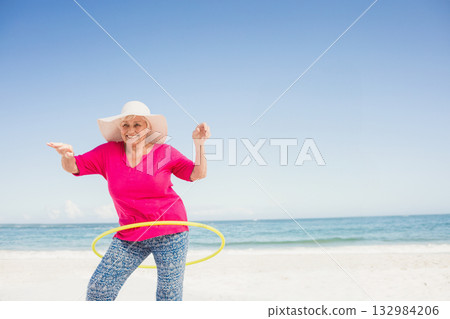 Senior woman in wide white sunhat spinning yellow hula hoop on sandy beach with turquoise ocean 132984206