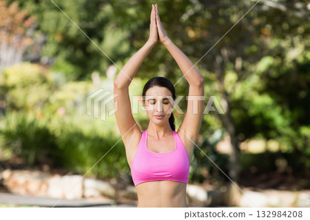Woman in 20s standing with arms raised overhead and palms pressed in garden wearing pink sportswear Woman in 20s standing with arms raised overhead and palms pressed in garden wearing pink sportswear 132984208